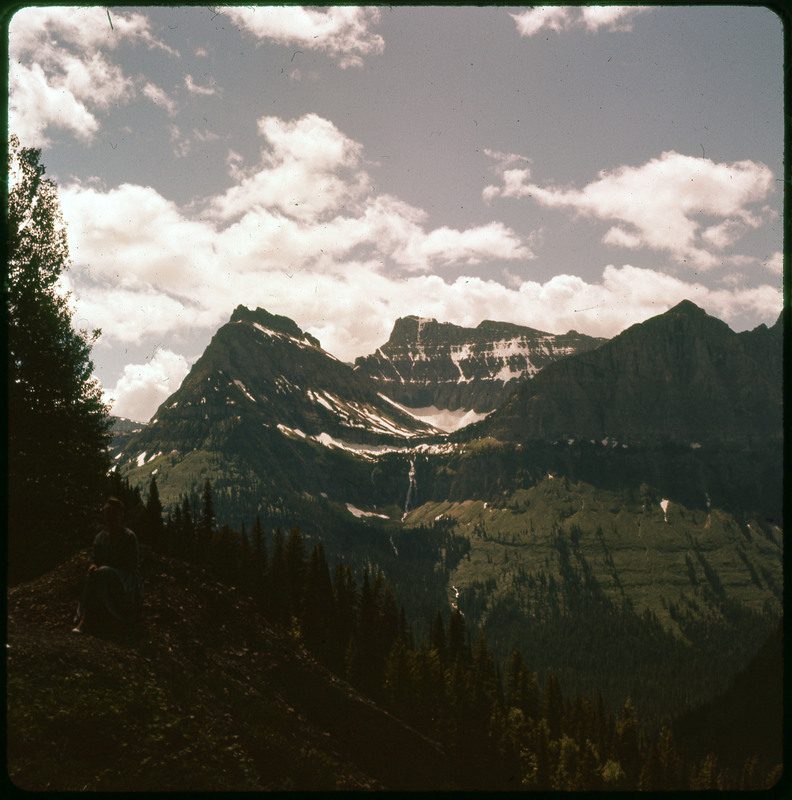 A photographic slide of a snowy mountain range with a waterfall, and many pine trees. There are clouds in the sky.