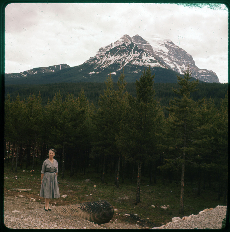 A photographic slide of Evelyn Crabtree in front of a large forest. There are snowy mountains in the background.