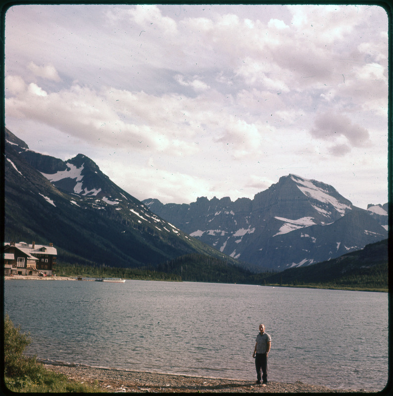 A photographic slide of a lake with a lodge on the shore. There are snowy mountains in the background.