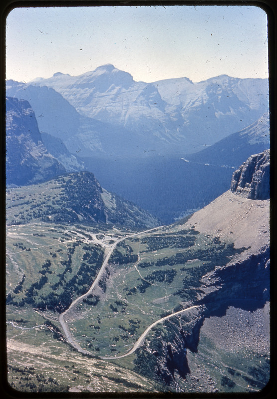 A photographic slide of a car driving on a road lined with a lot of snow pack. There are snowy mountains in the background.