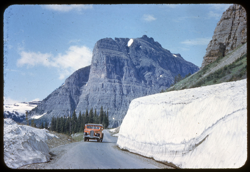 A photographic slide of a grassy and forested valley with many mountains in the background. There is a road through the valley.