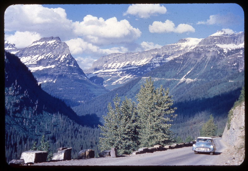 A photographic slide of a mountain range behind a car traveling down a road lined with high snow pack.