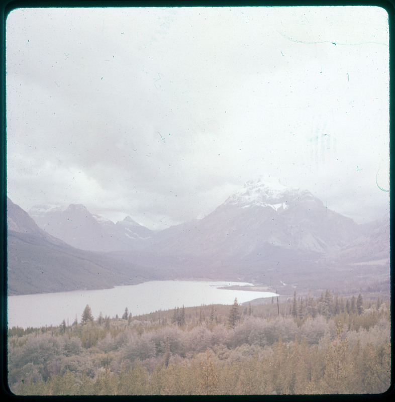 A photographic slide of a lake surrounded by forest. There are snowy mountains in the background and the sky is cloudy.