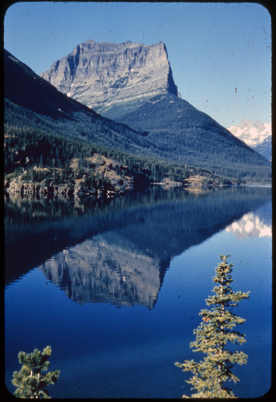 A photographic slide of a large rocky mountain and forest reflected in the surface of a lake.