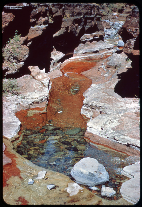 A photographic slide of a rocky stream in a rocky valley. The stream bed is red and the rocks are carved by the water.