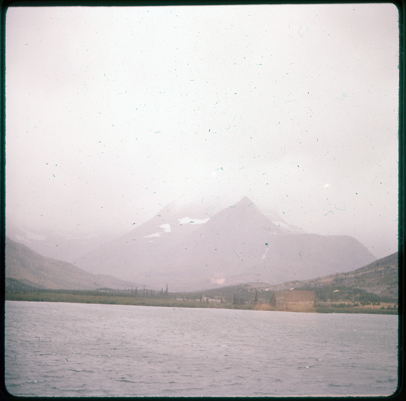 A photographic slide of a snowy mountain under a cloud. There is a lake in the foreground.