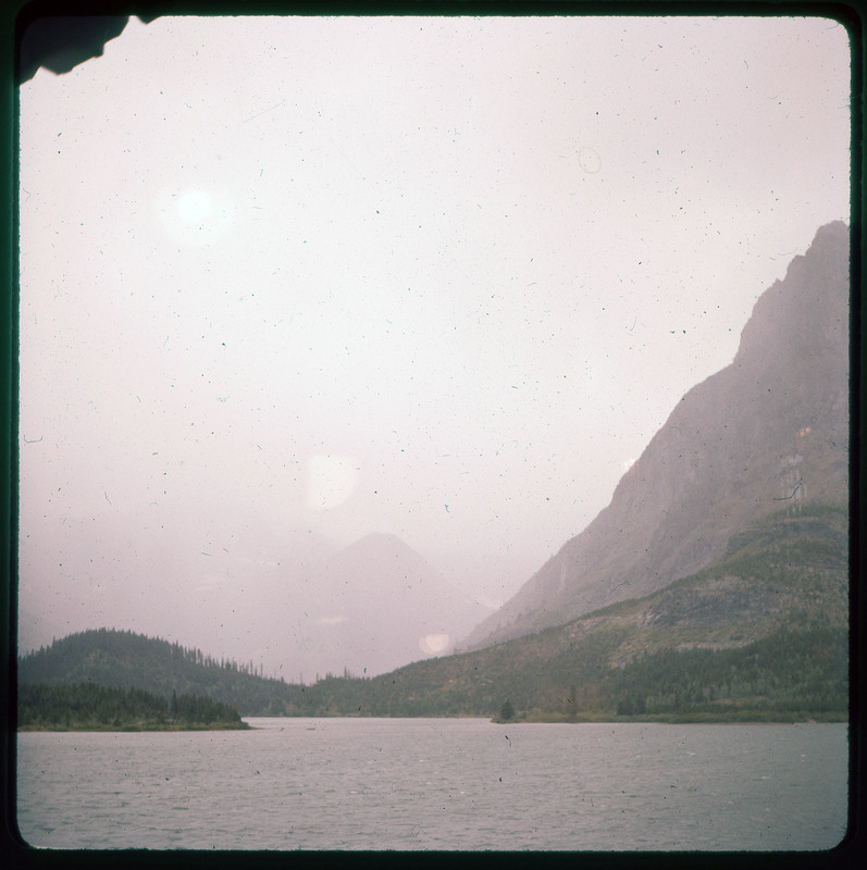 A photographic slide of a mountain valley. The mountains are either in fog, a cloud, smoke, or the image quality is poor. There is a forest in the foreground.