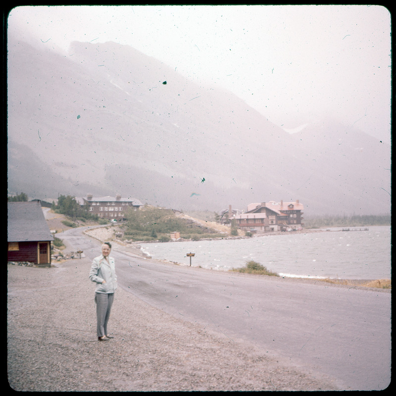 A photographic slide of Evelyn Crabtree standing beside a road. There are buildings in the background along a lake coast. There are foggy mountains in the background.