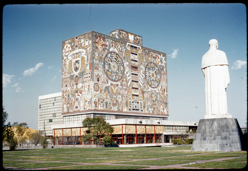 A photographic slide of a large building decorated with historical art next to a large statue of a person.