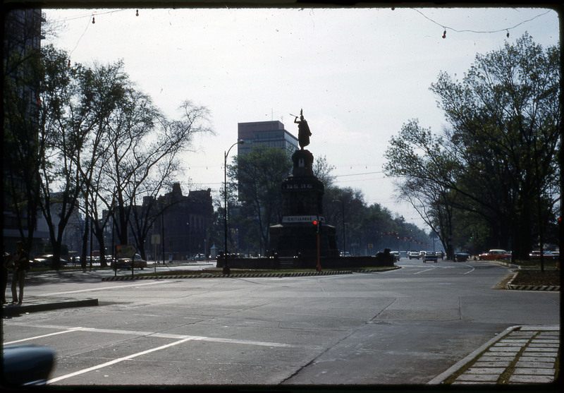 A photographic slide of a shaded statue of a warrior holding a spear in the middle of an intersection.