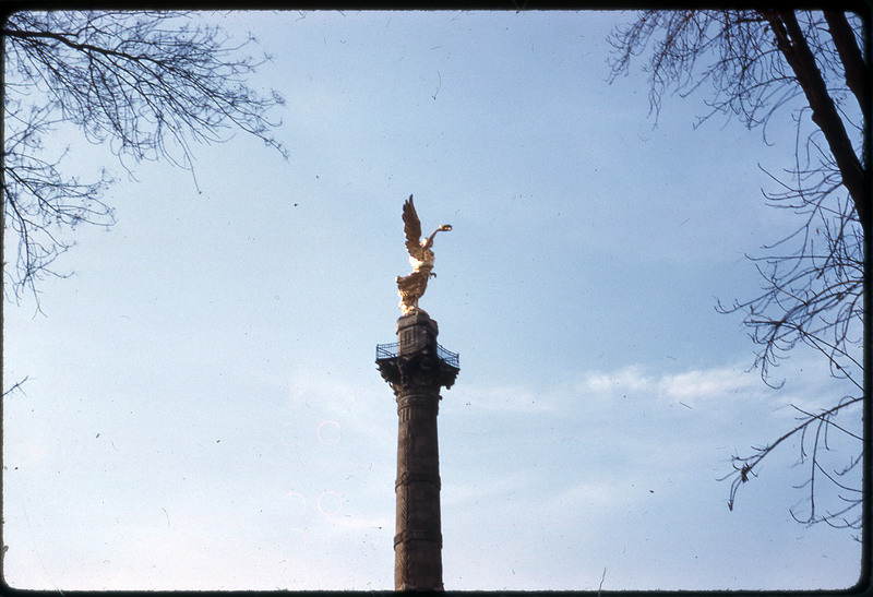 A photographic slide of a monument depecting a golden statue of an angel sitting atop an ornately carved column.