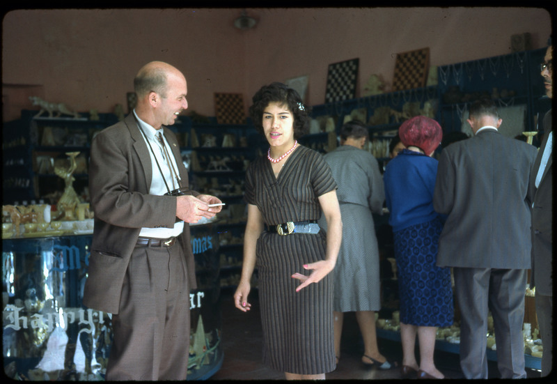 Photographic slide of a man and woman speaking in a storefront. There are other people in the background. Likely a shop in another country as there is writing in a different language.