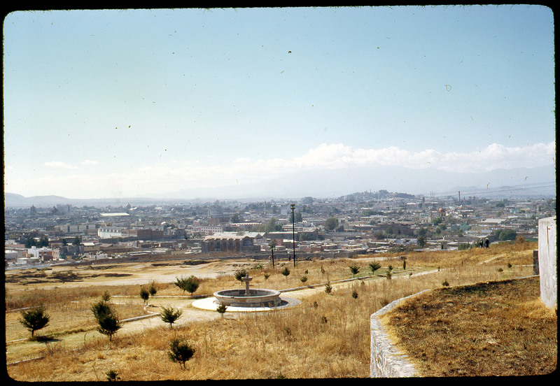 Photographic slide of a scenic view of a city from a high point. There is a water fountain and garden in the foreground with the city in the background.