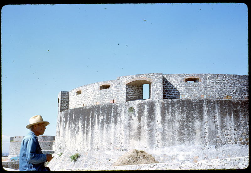 A photographic slide of a stone structure with windows, likely ancient. The foreground gestures a man in a hat.