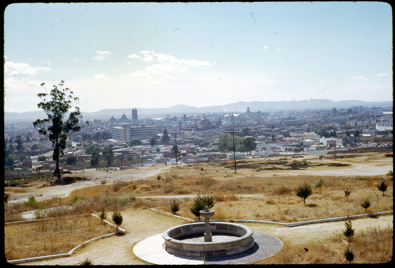 A photographic slide of a city in the background with mountains. In the foreground there is a garden and fountain.