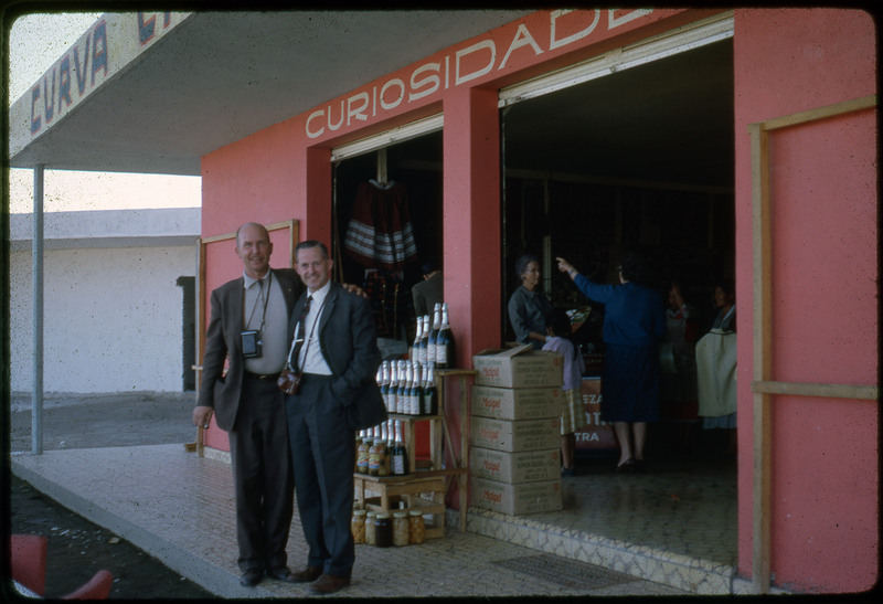 A photographic slide of Donald Crabtree posing with another man. They are outside a storefront, with people in the store and bottles of champagne out front.