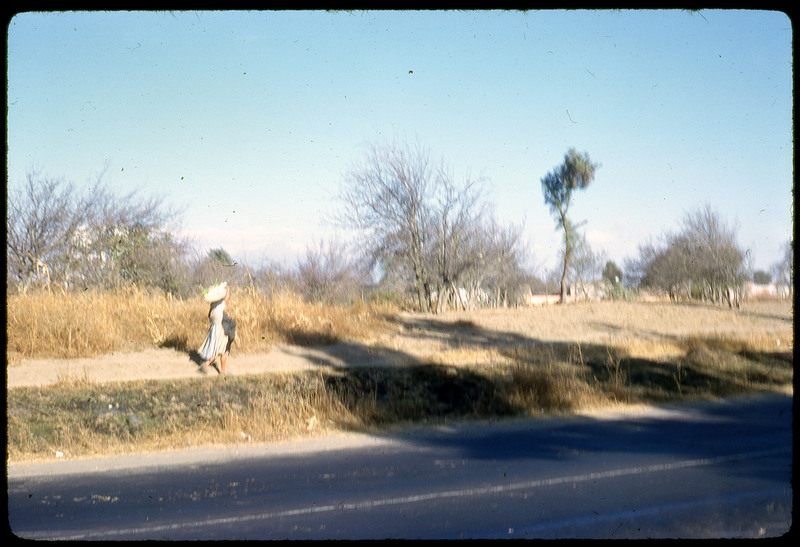 A photographic slide of a woman walking with a load of goods on her shoulder on a trail beside a road. The image is blurry and there are trees in the background.