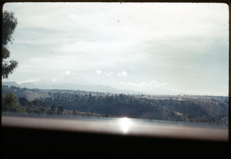 A photographic slide of a valley and mountain view. There are hills with trees in the foreground and faded mountains in the background.