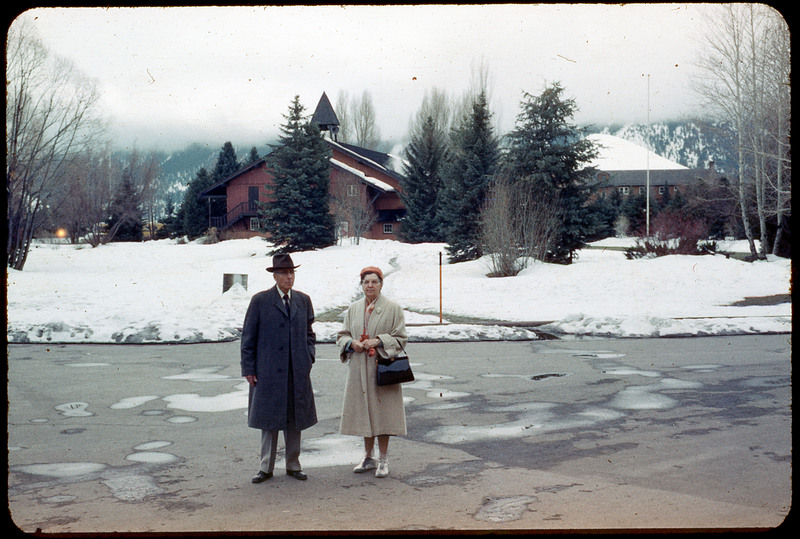 A photographic slide of a couple standing outside a house in a snowy landscape. There are pine trees and they are standing on the road.