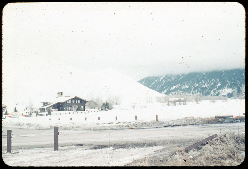 A photographic slide of a snowy landscape with a house. There is a snowy road and mountain that is in clouds.