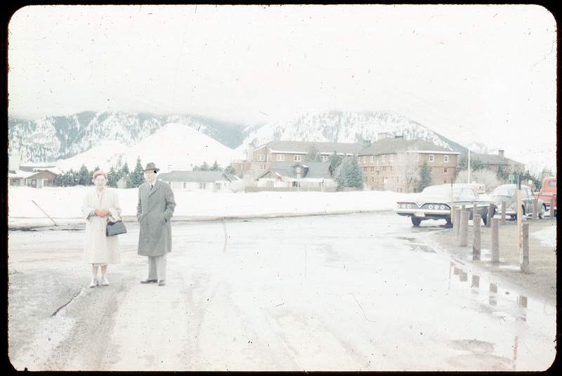 A photographic slide of a couple standing on the road in an snowy landscape. There are houses and mountains in the background.