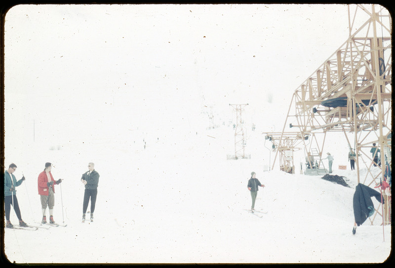 A photographic slide of people skiing and a chairlift. The image is mostly white, but there are five obvious skiers and some in the background.