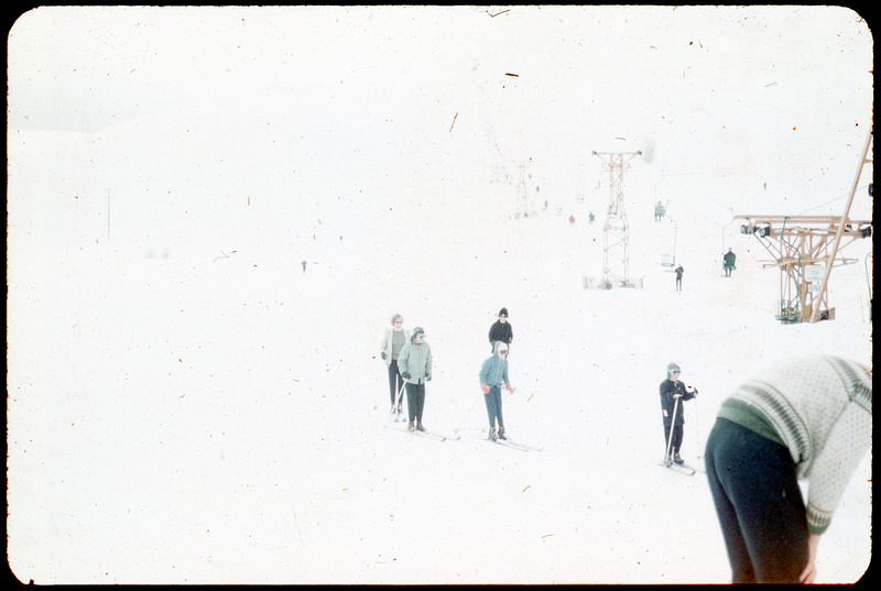 A photographic slide of people skiing and a chairlift. There five people actively skiing and you can see people sitting on the chairlift.