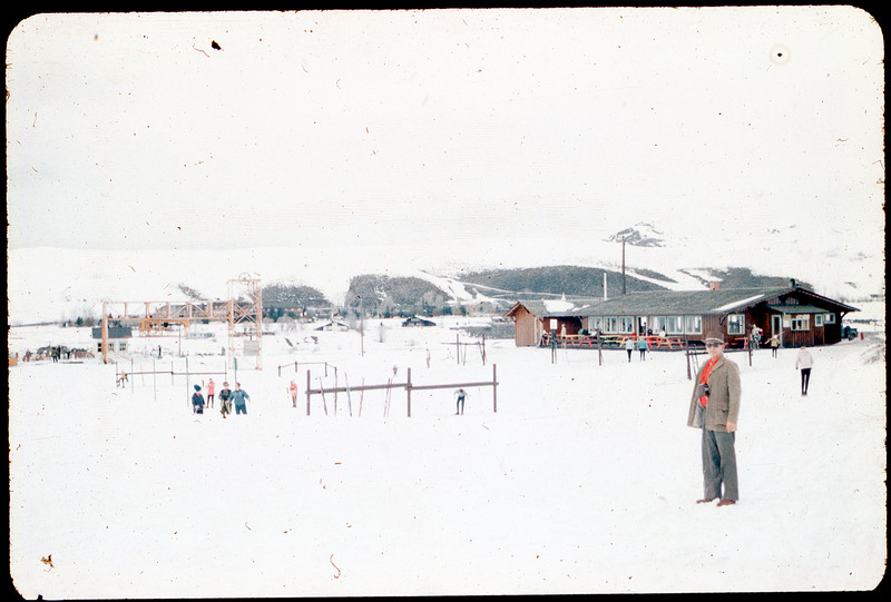 A photographic slide of Donald Crabtree standing in front of a ski resort. There are some people in the background and a ski lodge.