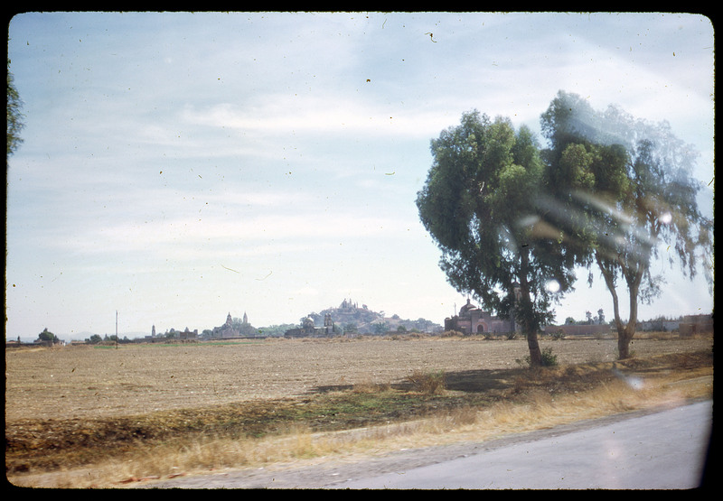 A photographic slide of a cityscape that seems to be lined with many temple-like buildings. The image is taken from the road and there is a field in the foreground with trees.