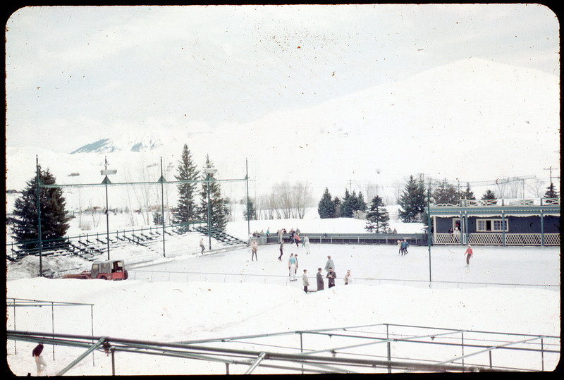 A photographic slide of an ice skating rink with many people there. There are snowy mountains in the background.