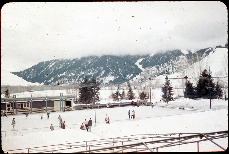 A photographic slide of an ice skating rink with many people skating and standing on the sides. There are snowy mountains in the background and a skating lodge.