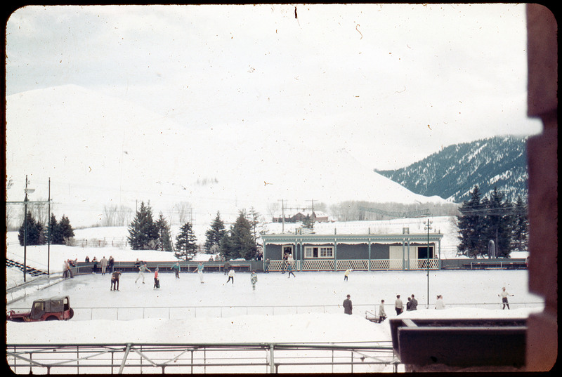 A photographic slide of an ice skating rink with many people skating. There are houses and snowy mountains in the background.