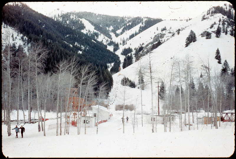 A photographic slide of a chair lift up a mountain. There are some people skiing in the foreground and aspen trees.