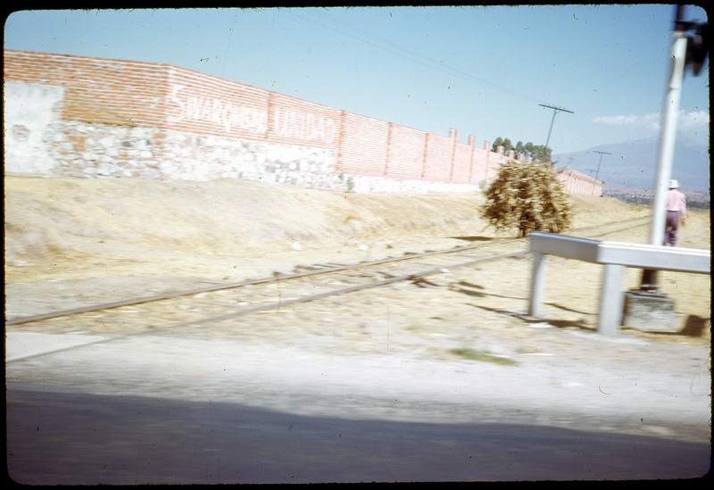 A photographic slide of a railroad and a fence with writing on it. There is a man on the side of the photo.