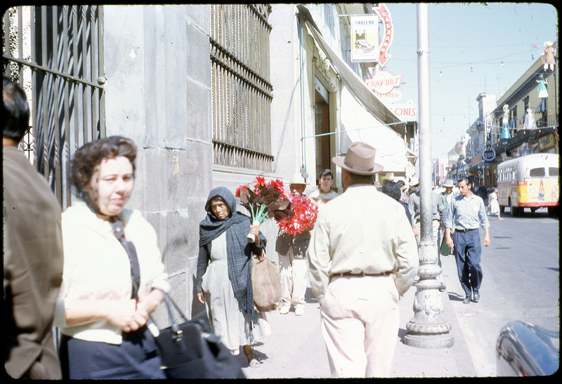 A photographic slide of people walking on the streets of a city. There is a bus in the background and many shops. This looks like it could be Mexico.