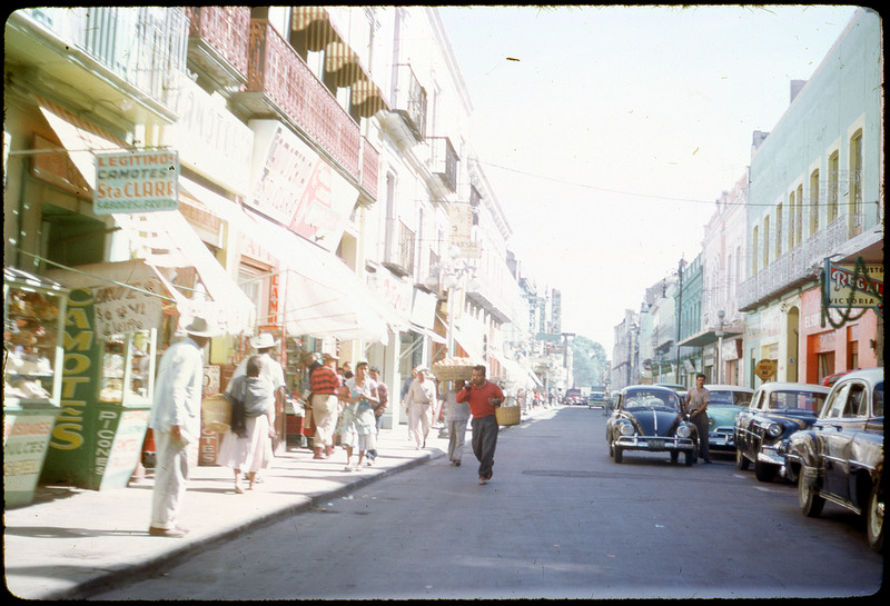 A photographic slide of people walking on a city street. There are cars parked on the side and people walking on the sidewalk. There are many shops with signs in Spanish.