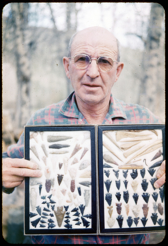 A photographic slide of a man holding a framed display of lithic tools. Most of the tools are arrowheads and bifaces.