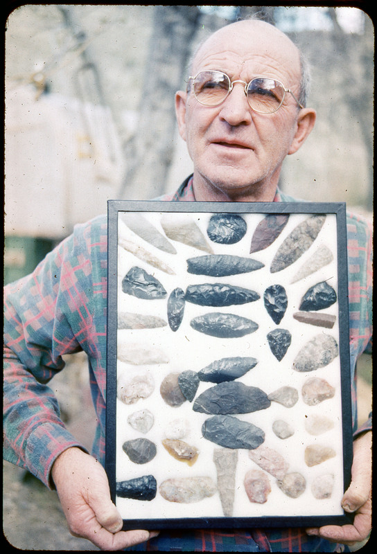 A photographic slide of a man holding a framed display of lithic tools. Some appear to be scrapers, but most are arrowheads. There is a tree and house in the background.