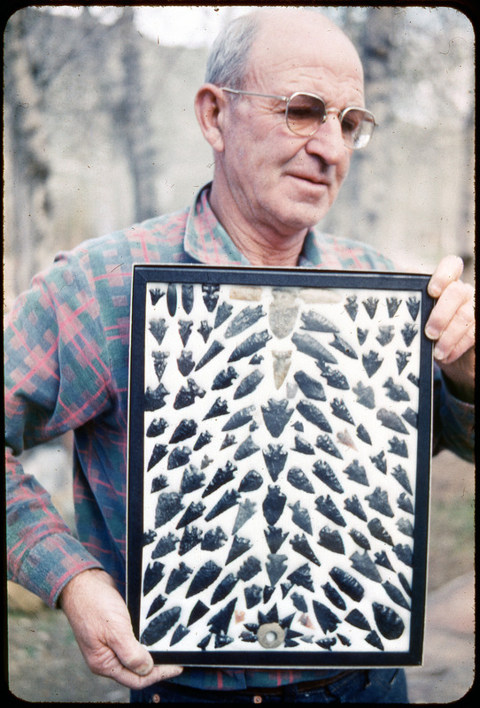 A photographic slide of a man holding a framed display of lithic tools. They all appear to be arrowheads.