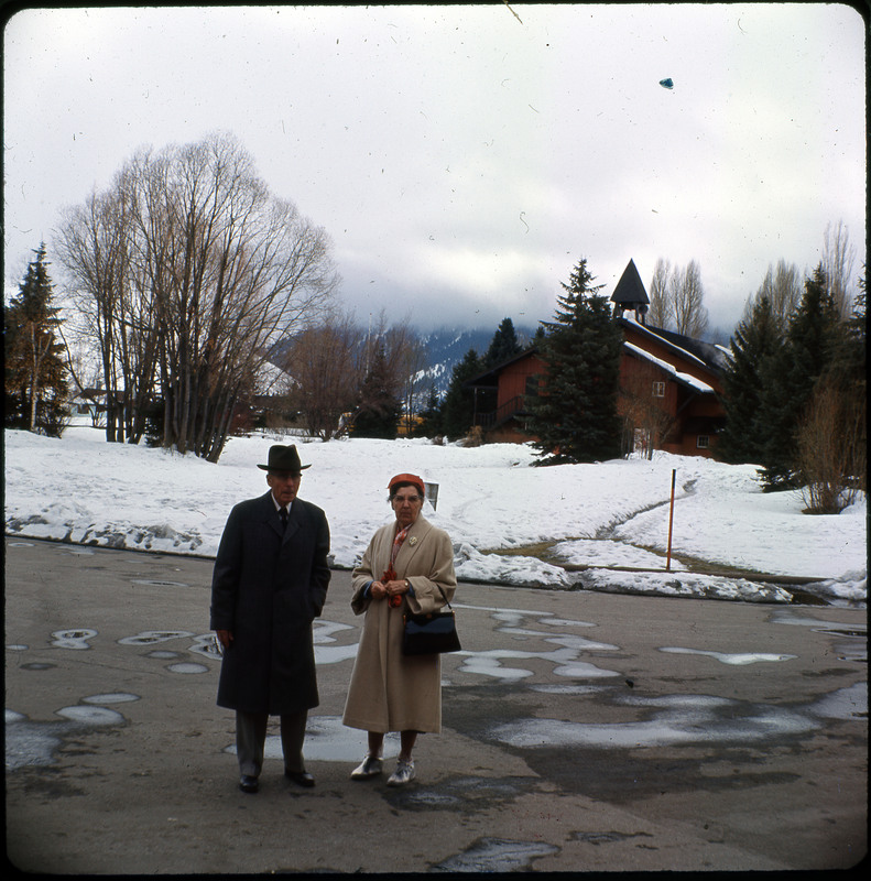 A photographic slide of a couple standing on a street in front of houses. There is a snowy lawn and mountain in the background.