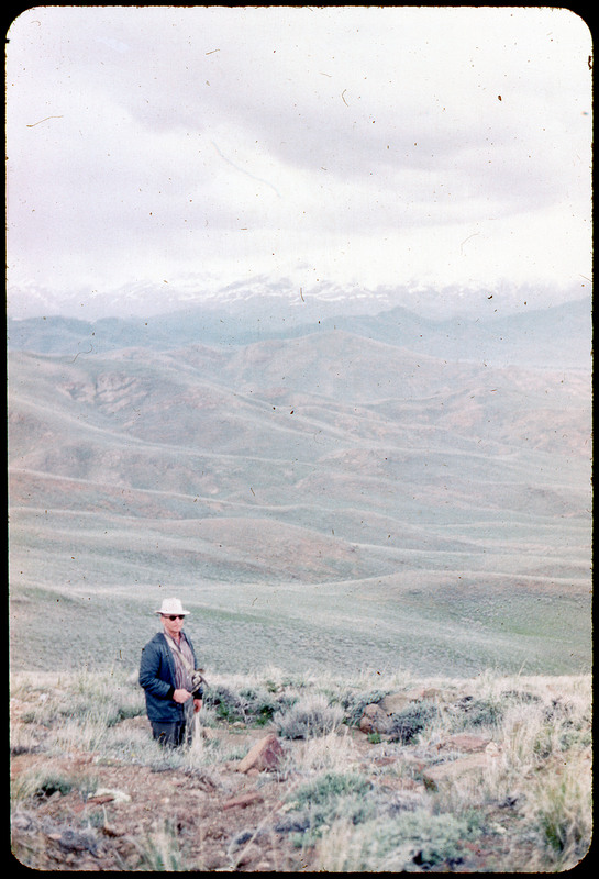 A photographic slide of Donald Crabtree standing on top of a hill. There is a snowy mountain range in the background and many grassy hills.