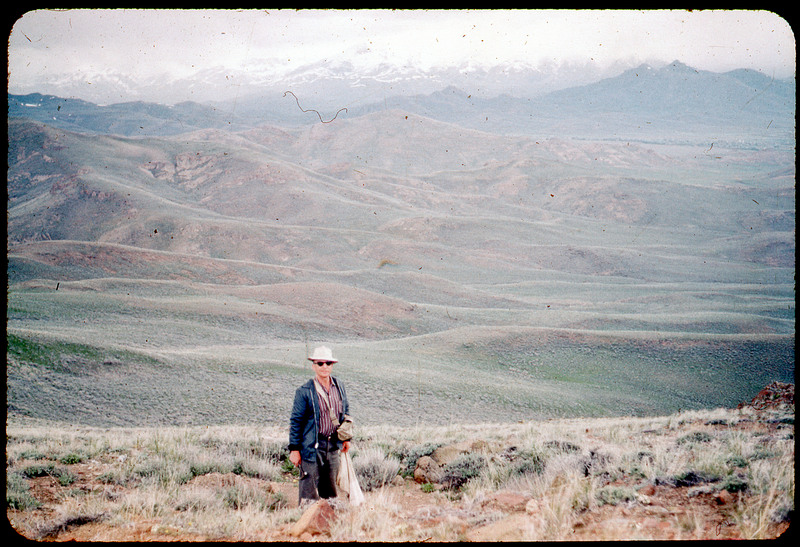 A photographic slide of Donald Crabtree standing on top of a hillside. He is holding what resembles a hammer. There are snowy mountains in the background and grassy hills.