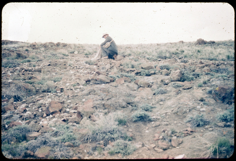 A photographic slide of Donald Crabtree collecting rocks on a grassy and rocky hillside. He is holding a bag of rocks.