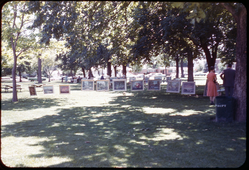 A photographic slide of paintings displayed for viewing in a park. There are people viewing the paintings.