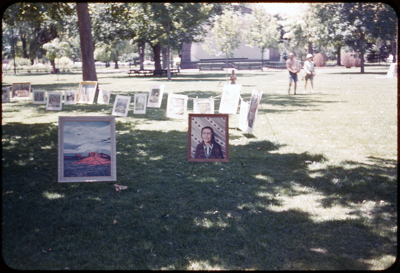 A photographic slide of paintings displayed in the park. There are two paintings in the foreground, one of a woman and one of a landscape. There are other paintings in the background and people in the park.