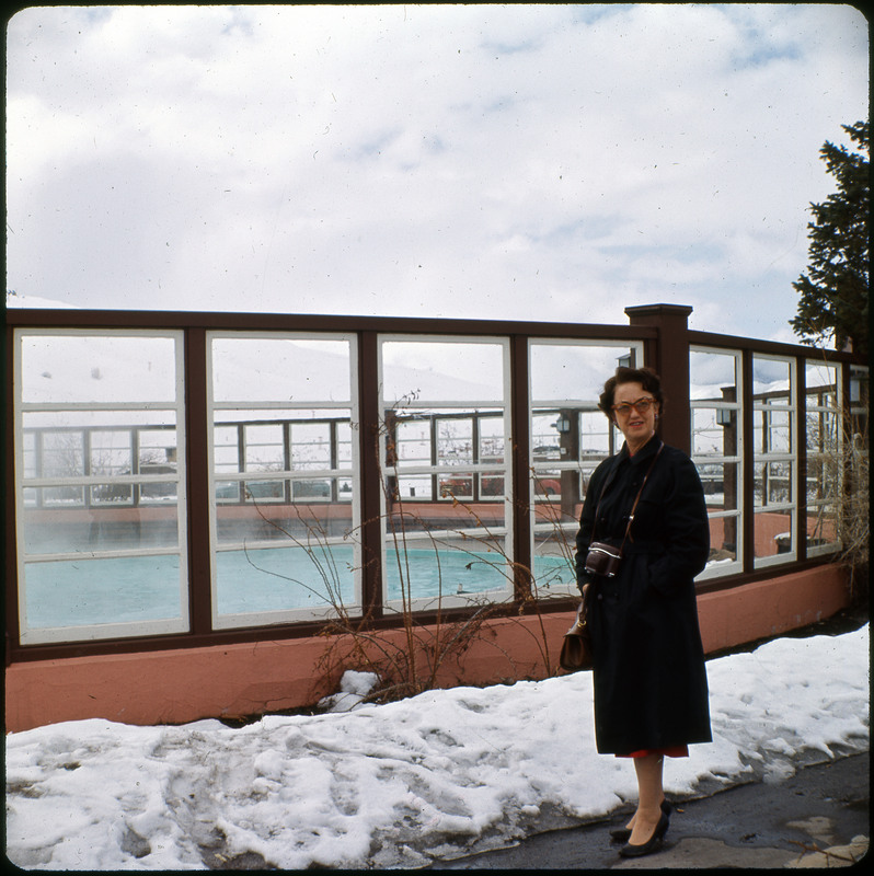 A photographic slide of Evelyn Crabtree standing in front of a hot springs. The pool is enclosed and there is snow on the ground.