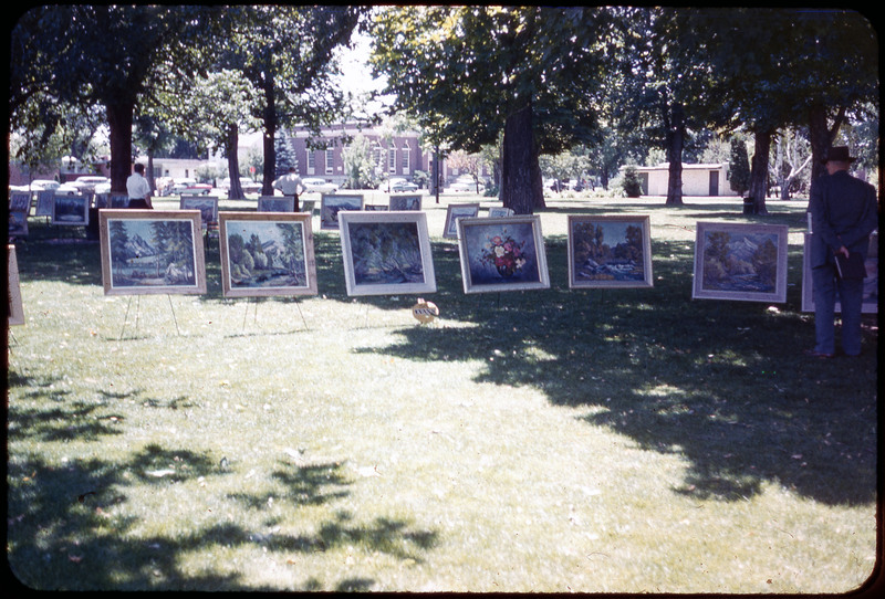 A photographic slide of paintings displayed in a park. There are many paintings of landscapes and people in the background.