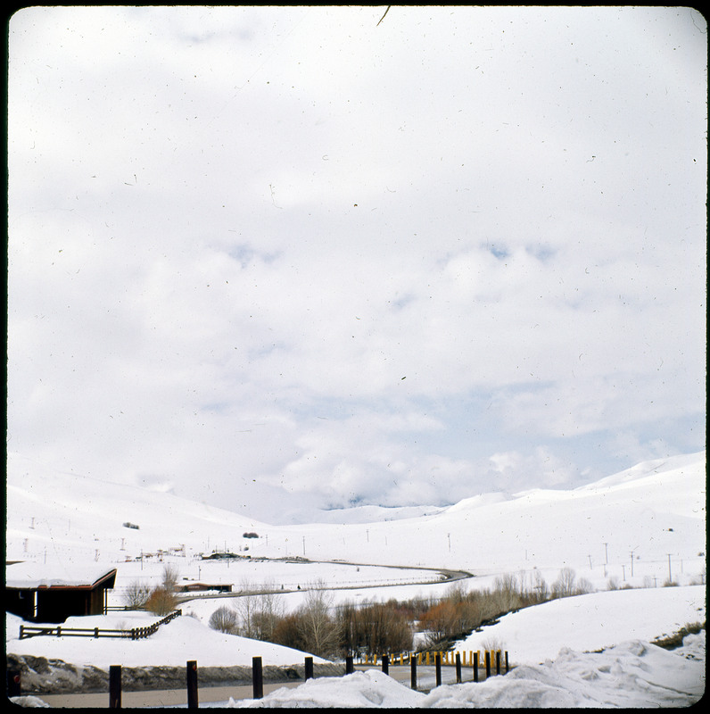 A photographic slide of a snowy mountainous landscape. There is a road and power lines in the photo, and a very cloudy sky.