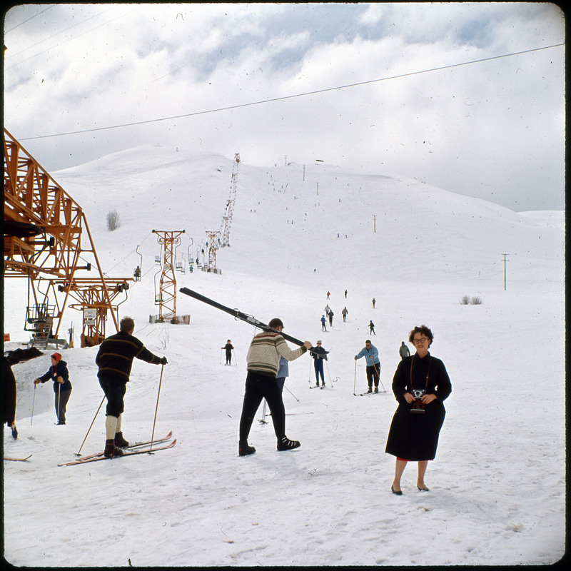 A photographic slide of a ski resort. There is a chairlift in the photo and many skiers. Evelyn Crabtree is posing in front of the hill.