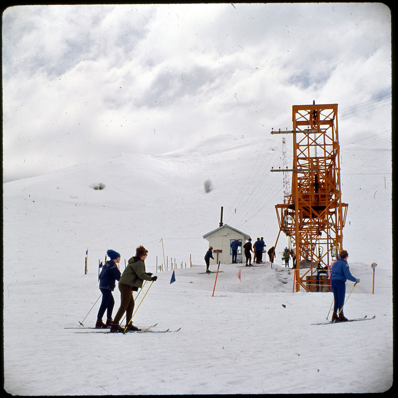 A photographic slide of a ski resort. There is a chairlift and many skiers in line at the lift. It is a snowy landscape.